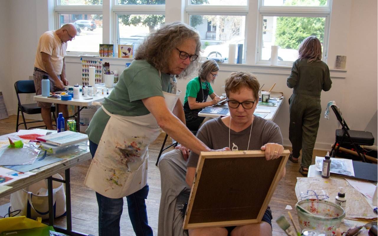 A spacious art studio filled with natural light. In the foreground, a teacher assists a seated student, who is holding a canvas, while another student works at a table in the background. Various art supplies, including paints and brushes, are scattered around the room, and additional students are seen engaged in their artwork.