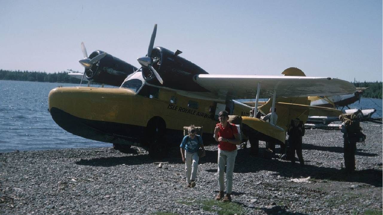 A bright yellow and black seaplane, identified as Isle Royale Airways, is partially on a rocky shore next to a lake. Two individuals are walking away from the plane, one is a child in a blue shirt and the other an adult in a red shirt and light pants. In the background, other seaplanes are visible on the water, surrounded by dense green trees under a clear blue sky.