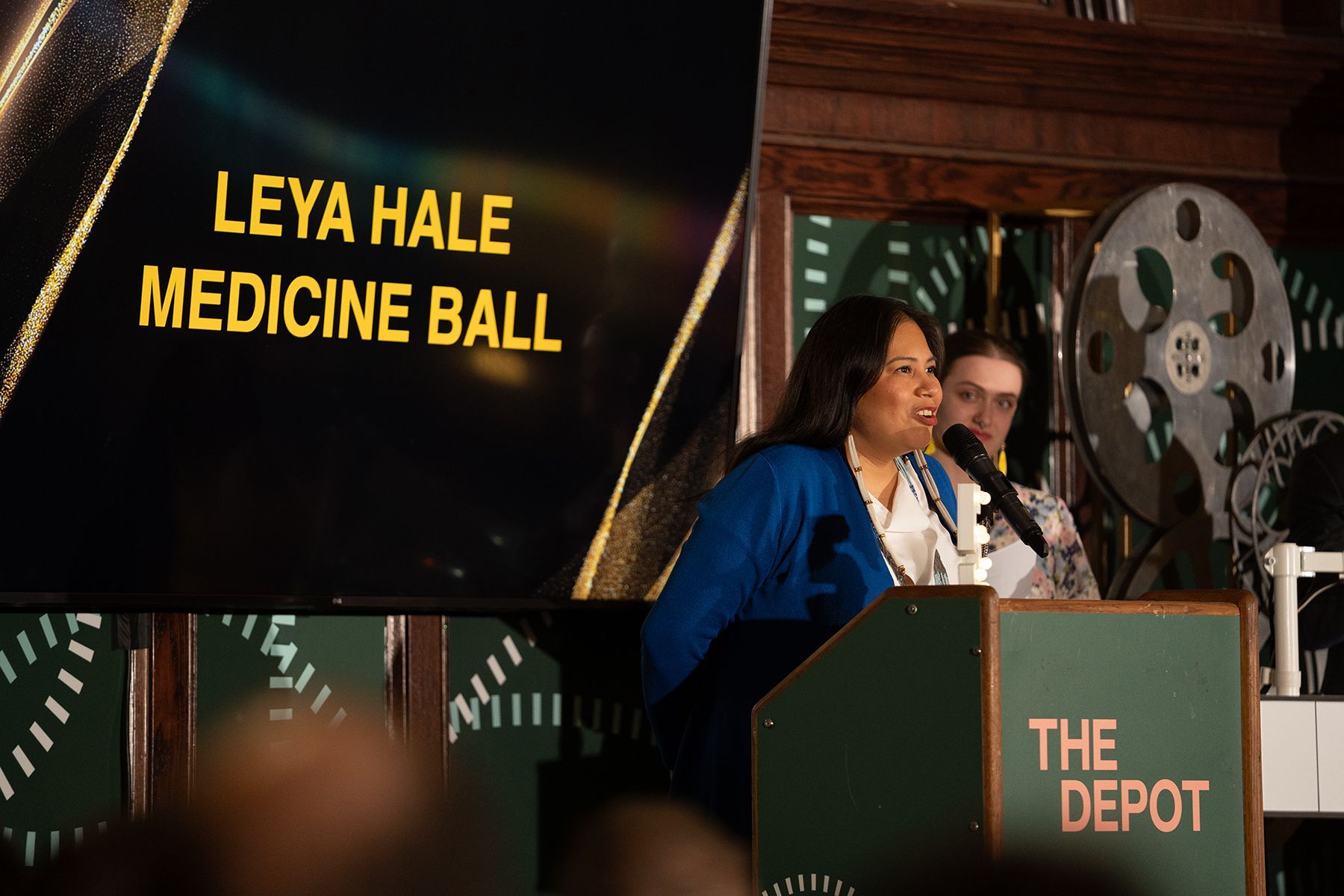 A woman wearing a blue blazer and white shirt speaks at a podium labeled "THE DEPOT" with a microphone. Behind her is a large screen displaying the text "LEYA HALE MEDICINE BALL." Another person stands in the background near a decorative film reel.