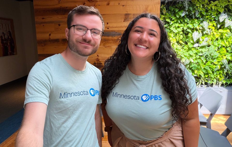 Two people smiling and posing together indoors. Both are wearing light blue t-shirts with the "Minnesota PBS" logo. The background features a wooden wall and a green leafy plant display.