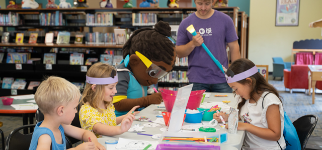 Children engaged in a craft activity at a table in a library, accompanied by a person in a large mascot costume and a staff member holding a large foam brush.