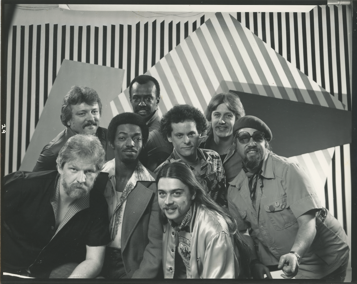 Black-and-white photo of a group of eight men posing closely together in front of a geometric, striped background. The men display a variety of styles including casual and patterned shirts, with one wearing a beret and sunglasses.