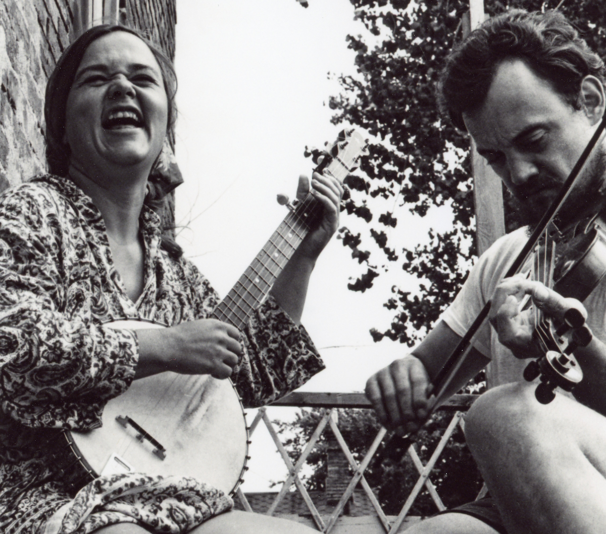 Black-and-white photo of a woman smiling broadly while playing a banjo, sitting next to a man focused on playing a violin; they are seated on a porch with a tree and latticework in the background.
