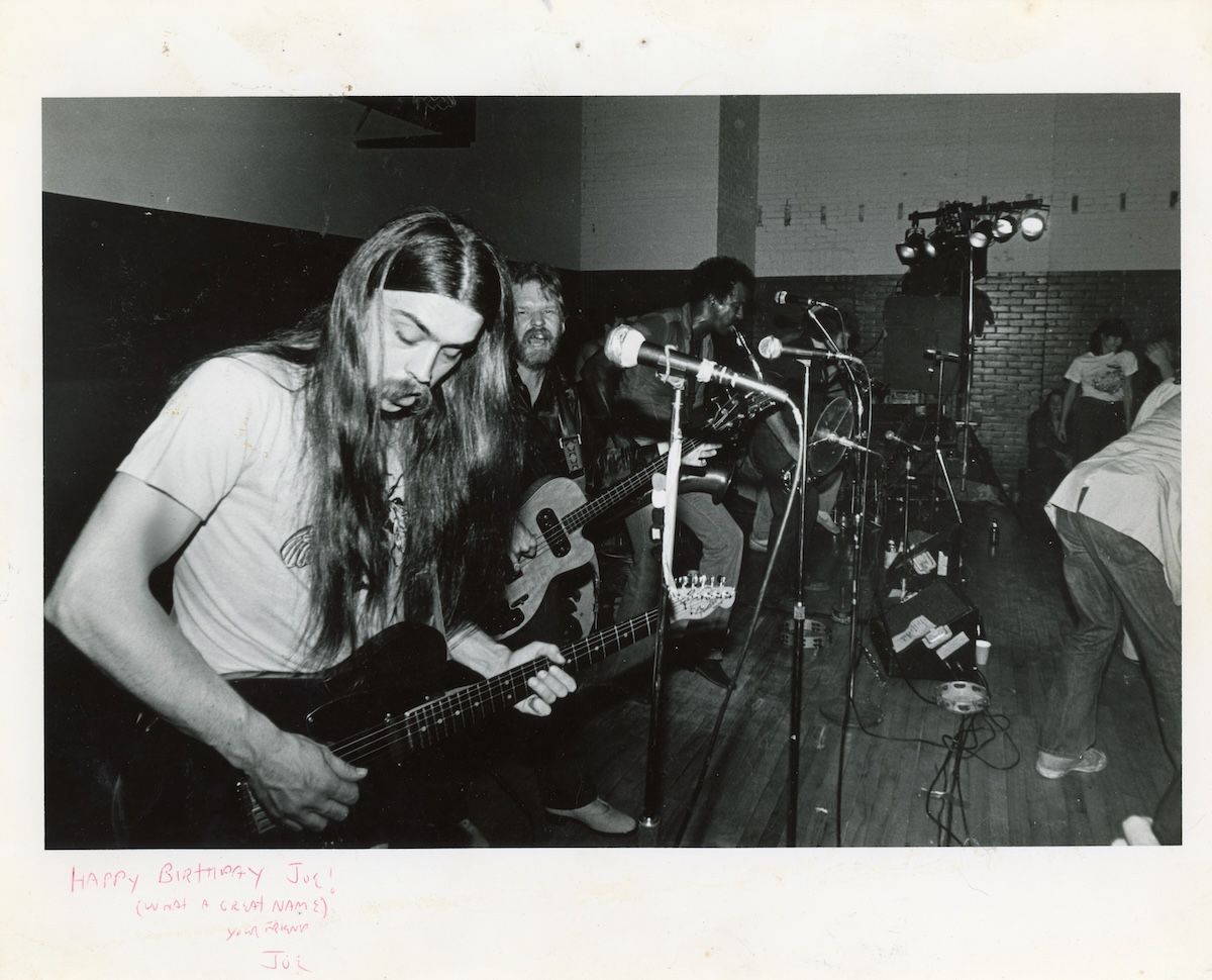 Black-and-white photo of a band performing on stage, featuring a long-haired guitarist in the foreground playing an electric guitar, with other band members playing bass and saxophone in the background, and stage lights and microphones visible.