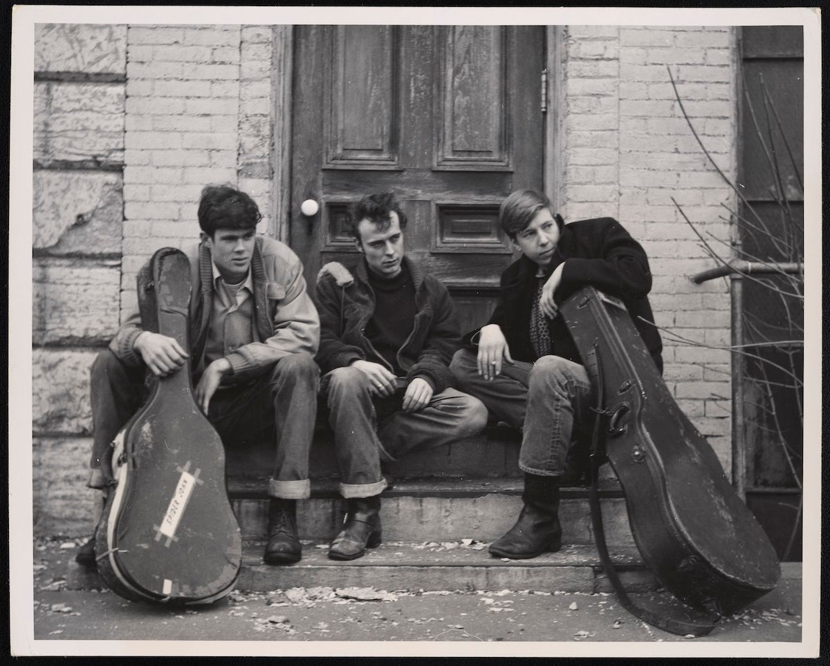 Black and white photo of three young men sitting on stone steps in front of a wooden door, each with a guitar case. The two men on the left and right are holding their guitar cases upright, while the middle man sits with hands clasped. The surrounding walls are made of brick and stone.