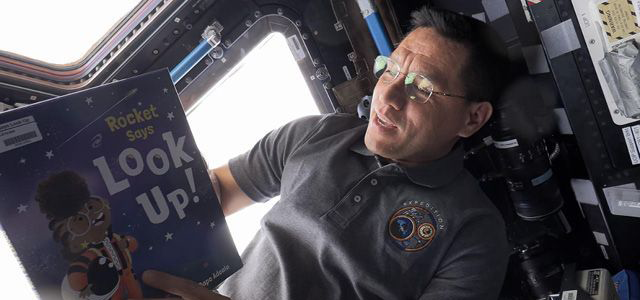 Astronaut inside a spacecraft reading the children's book "Rocket Says Look Up!" near a window with sunlight streaming in.