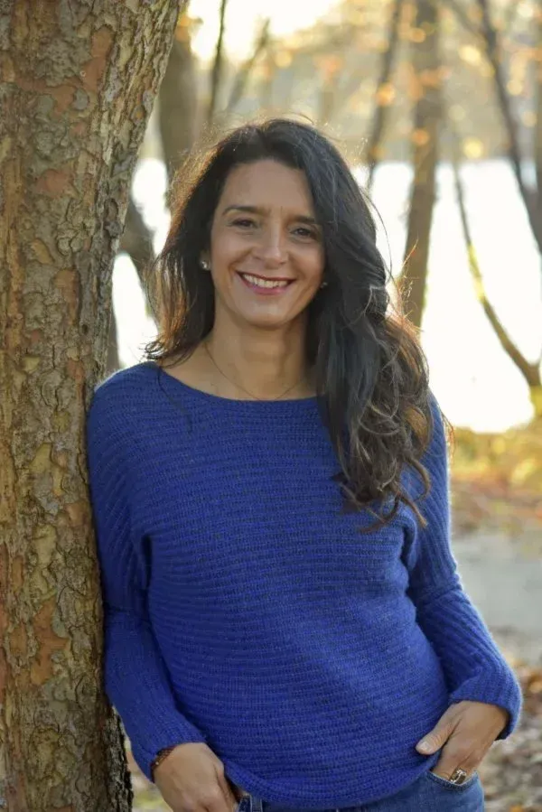 A woman with long, wavy dark hair smiles while leaning against a tree. She wears a blue sweater and is standing near a calm body of water, surrounded by autumn foliage. The sun filters through the trees, creating a warm atmosphere.