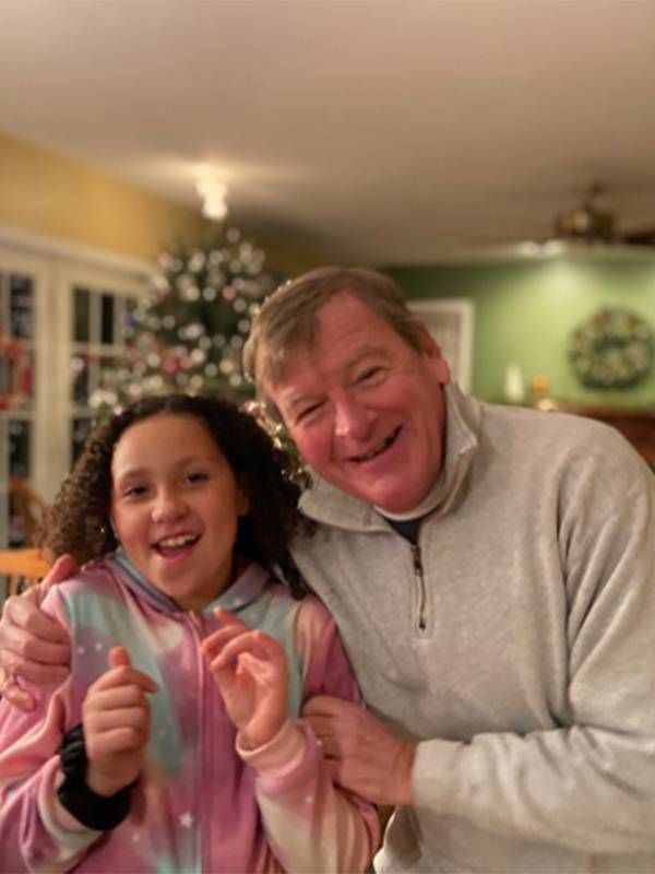 A smiling man in a light gray quarter-zip sweater embraces a cheerful young girl wearing a colorful hoodie, with a decorated Christmas tree and holiday wreath in the background.
