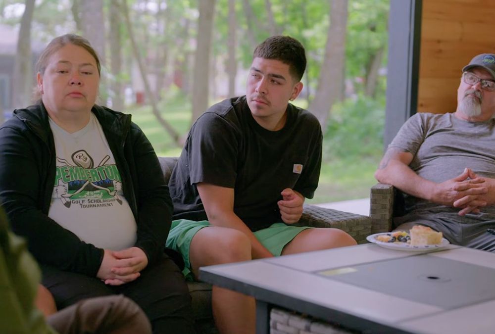 Three people sit on a couch outdoors: a woman in a black jacket and white T-shirt, a young man in a black T-shirt and green shorts, and an older man in a gray T-shirt and baseball cap. A plate with food is on the table in front of them.