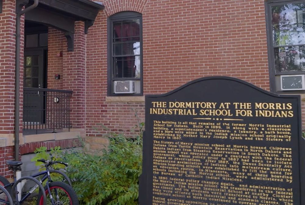 A brick building with two windows and a black door with a porch. In front of it, a black sign with gold text titled "The Dormitory at the Morris Industrial School for Indians," providing historical information. A bicycle is parked next to some green plants near the building.