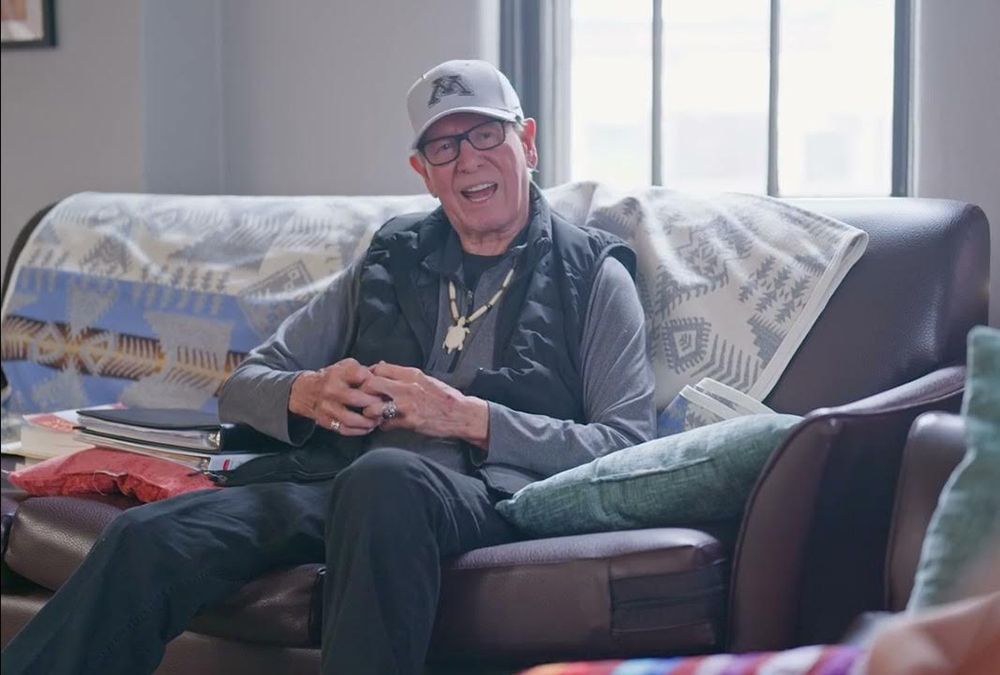 Older man wearing a gray baseball cap, glasses, and a black vest sitting on a brown leather couch with patterned blankets and cushions, smiling and gesturing with his hands in a well-lit room.