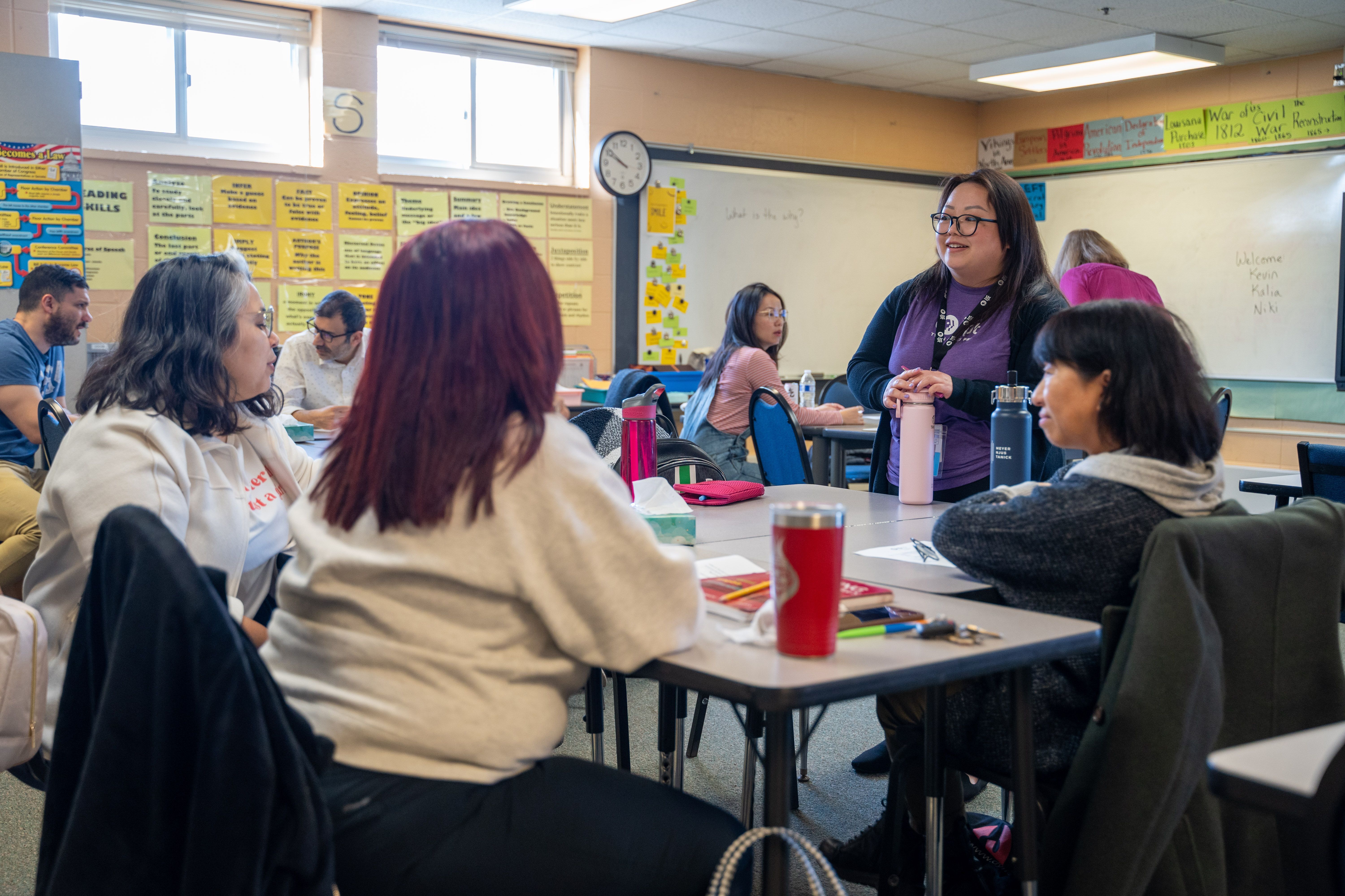 A diverse group of adults engaged in a classroom discussion, with three women sitting at a table in the foreground, one standing and speaking, and other participants seated at desks in the background. The classroom has educational posters and a whiteboard with writing.