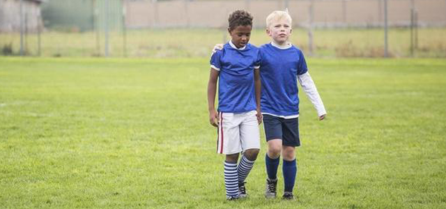 Two young boys wearing blue soccer jerseys walking on a grassy field, one boy has his arm around the other.